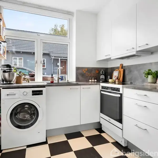 Et køkken med hvide skabe og sorte håndtag. En vaskemaskine og blender står på bordpladen ved et vindue. Skakternet sort-hvidt gulv og krydderihylder på væggen.