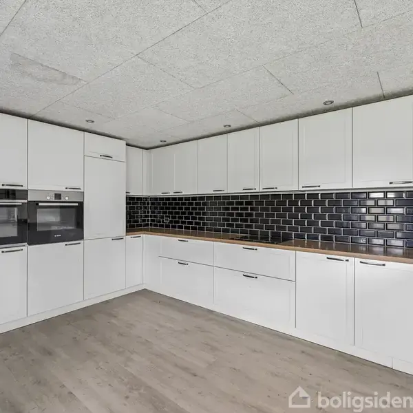 Et moderne køkken med hvide skabe omslutter et sort flisebelagt vægpanel. Trægulve understøtter det minimalistiske design, og indbyggede ovne er placeret til venstre. Ingen tekst i billedet.