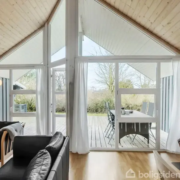 Stuen har sorte sofaer og et trægulv foran store vinduer med hvide gardiner, der åbner ud til en terrasse med havemøbler i en grøn, frodig have.