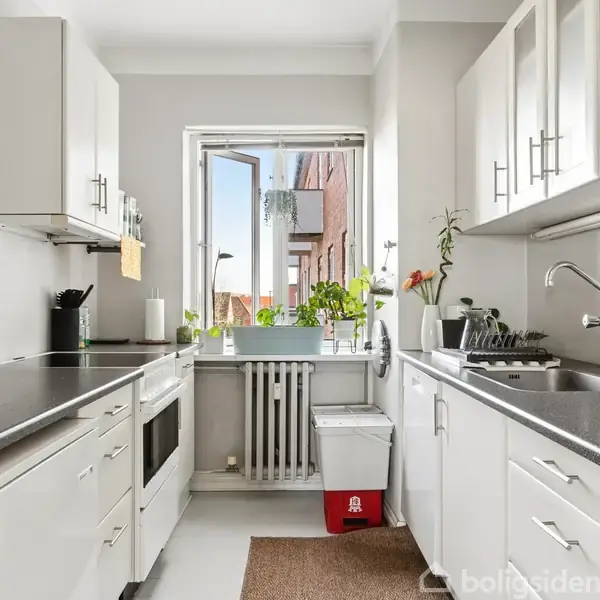 Et funktionelt køkken med hvide skabe og sorte bordplader. Vindue åbent mod en bygning udenfor, blomsterkrukke på vindueskarmen. Rent og sollyst miljø. Skilt bagved viser tekst: "Boligsiden".