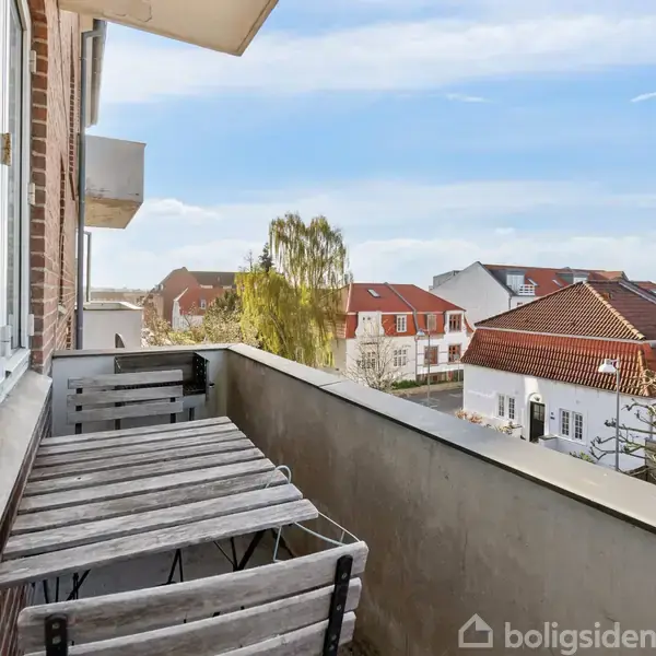 Balkon med træbord og stole placeret mod gelænderet, omkranset af udsigt til rødtagsboliger og træer. Skilt i vinduet med teksten "TIL SALG".