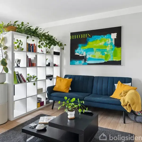 Reol med planter og bøger står ved siden af en blå sofa med gule puder. Et farverigt maleri hænger over sofaen i en lys stue med trægulve.