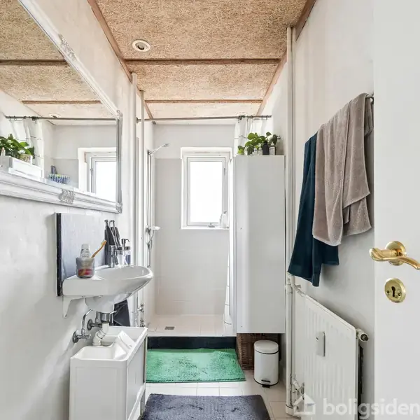 Håndvask med spejl hænger på en hvid væg, omgivet af planter og håndklæder. Badeværelset har et vindue og bruser bagved, med et lyst loft.