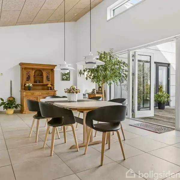 Et moderne spisested med et rundt træbord omgivet af fem sorte stole. Store vinduer og døre åbner ud til en terrasse. Planter og en kommode pynter rummet.