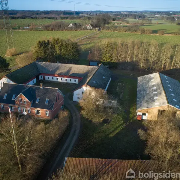 En firkantet gårdkompleks med rødt og hvidt murværk og et stort, gråt tag. Det ligger i et landskab med åbne marker og spredte træer under en klar himmel.