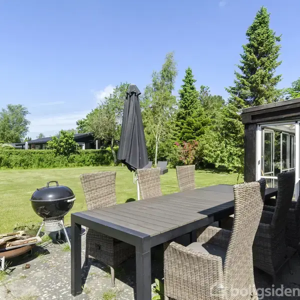 Terrasse med spisebord i flet og seks stole står under en sort parasol. En kuglegrill og pejs er til venstre. Området er omgivet af en grøn have og buske.