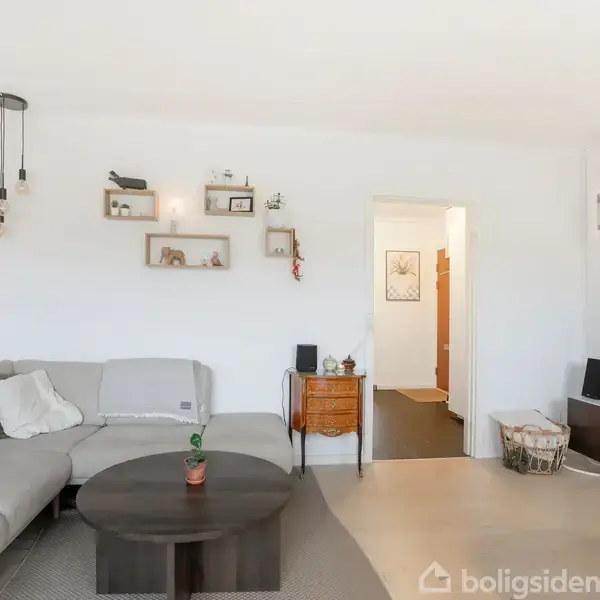 En stue med en grå hjørnesofa, sofabord og væghylder med dekorationer. Fjernsyn står på et skab, og der er adgang via en dør til en anden del af boligen.
