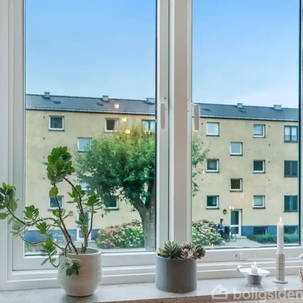 En vindueskarm med planter og stearinlys. Udenfor ses en gul boligbygning og træer under en blå himmel.