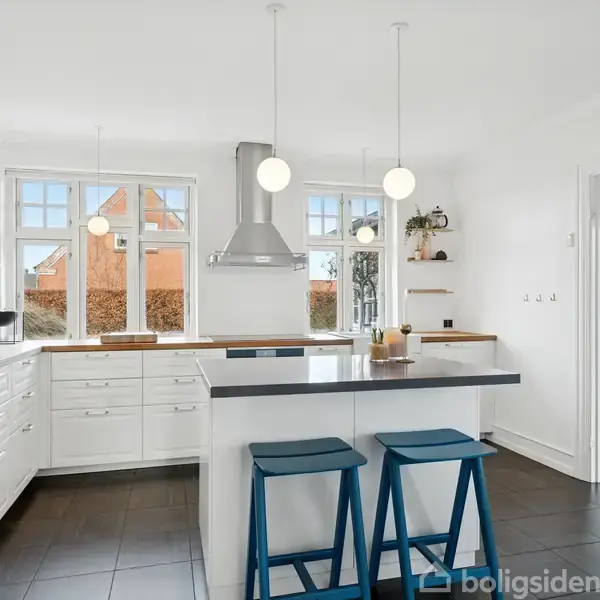Et lyst køkken med hvide skabe og en grå køkkenø. To blå barstole står ved øen. Tre kugleformede lamper hænger fra loftet. Vinduerne viser en udsigt med et rødt hus.