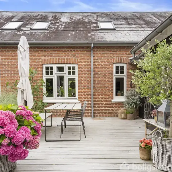 Terrasse med bord og stole, omgivet af blomster i kurve. I baggrunden ses en murstensbygning med hvide vinduer og ovenlysvinduer. Tæt ved bordet står en hvid parasol.
