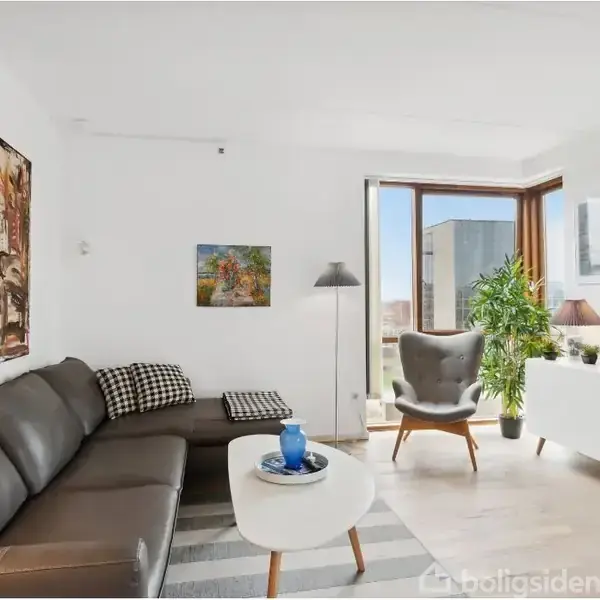 En moderne stue med en grå lædersofa står mod en hvid væg med abstrakt kunst. Et rundt bord har en blå vase. Store vinduer giver udsigt udenfor.