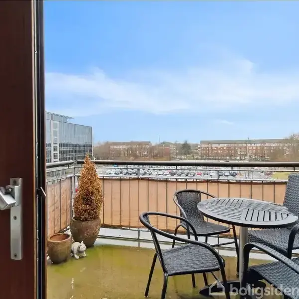 En åben altandør fører til en terrasse med et rundt bord og fire stole. Et par planter står ved rækværket, og i baggrunden ses en parkeringsplads og bygninger.