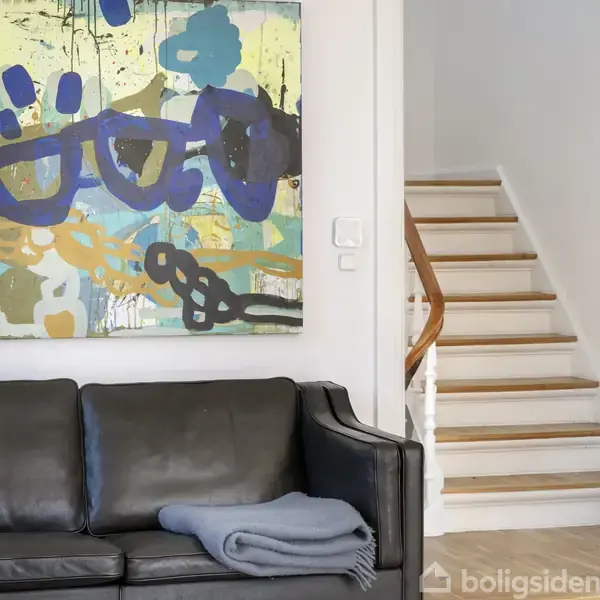 Sort lædersofa står foran en hvid væg med farverigt abstrakt maleri. Ved siden af sofaen er en trætrappe med hvidt gelænder, der fører opad.