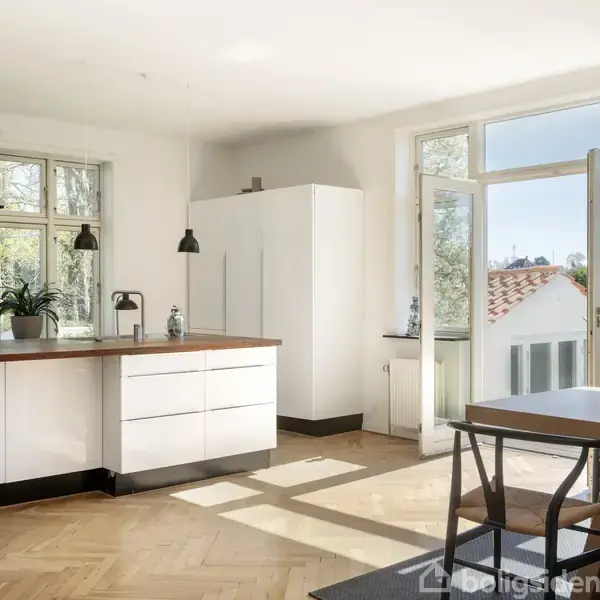 Et lyst køkken med hvide skabe og en træbordplade. Sollys strømmer ind gennem store vinduer og en altandør. En plante står på bordet, og en lampe hænger over køkkenområdet.