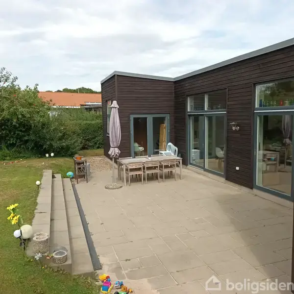 Flisebelagt terrasse med træbord og stole, omkranset af brun moderne bygning med store vinduer; baggrunden har grønne buske og en græsplæne. En parasol står ved bordet.