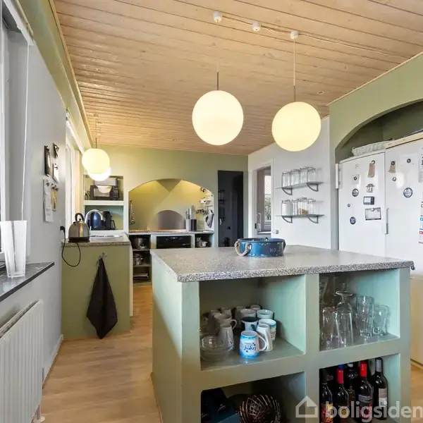 Køkken med en stor, lys køkkenø fyldt med glas og kopper; hængende runde lamper ovenfor. Køleskab med magneter til højre, træloft og store vinduer til venstre.