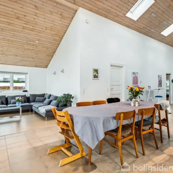 Et spisebord med træstole står i et rummeligt, lyst værelse med loft til kip. Til venstre er en sofagruppe, og baggrunden har planter og dekorative elementer.