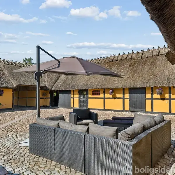 Gruppe af udendørs loungemøbler står under en stor parasol på en brostensbelagt gårdsplads. Rundt om er der gule bygninger med stråtage i en landlig setting.