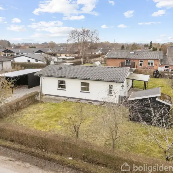 Et hvidt enfamilieshus står på en græsplæne med en hæk omkring. Ved siden af huset er en carport. Baggrunden viser en række huse under en blå himmel med skyer.