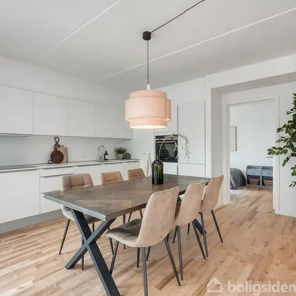 Et spisebord med seks stole står i et lyst køkken. Bordet har en enkel sort lampe over sig. Til højre ses en stor gulvplante. Væggene er hvide.