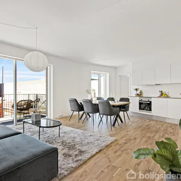 En moderne stue med åben køkken. En grå sofa står ved et sofabord. Spisebord med seks stole i centrum. Store vinduer giver udsigt til en balkon.