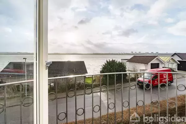 Udsigt fra altan over en vej med en rød postbil parkeret. I baggrunden ses huse og en flod under en overskyet himmel.