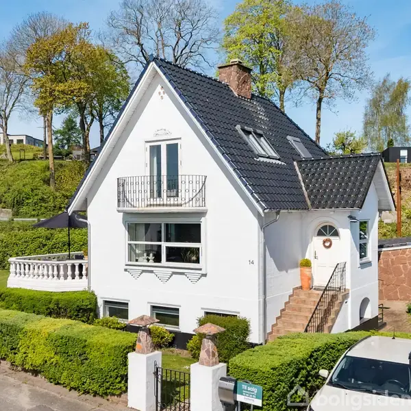 Et hvidt hus med sort tag står bag en hæk. Der er en trappe til hoveddøren og en balkon. En bil er parkeret ved indkørslen.