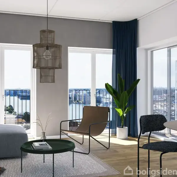 Stue med moderne møbler, herunder en grå sofa og et rundt bord, står foran store vinduer, der giver udsigt over en by og vand. Belysning hænger fra loftet.