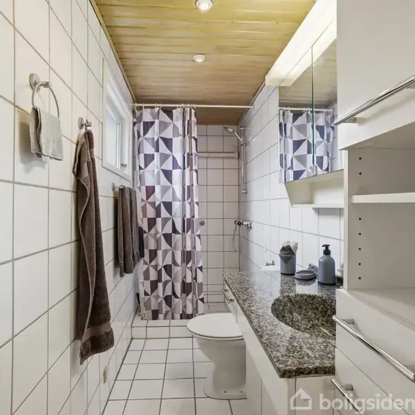 Badeværelse med hvidt flisegulv og vægge. Toilet og vask med spejl til højre. Geometrisk mønstret bruseforhæng for enden. Håndklæder hænger på væggen. Reol til højre.