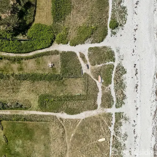 Strandområde set oppefra med græsarealer og stier før en strand med rullesten. To huse er synlige øverst og nederst til venstre med grønne buske imellem.