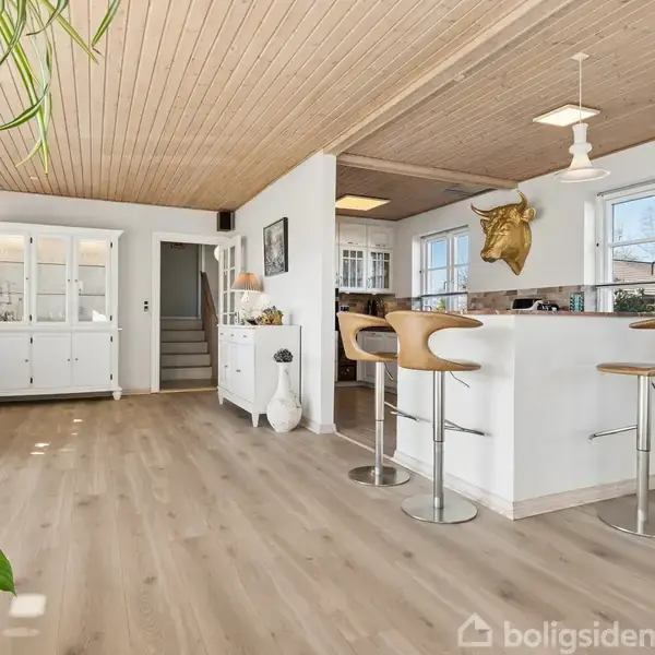 Et moderne køkken med en hvid bar, omgivet af træbarstole, står i et rummeligt værelse med træloft og gulv. Grønne planter tilføjer livlighed i det lyse rum med store vinduer.