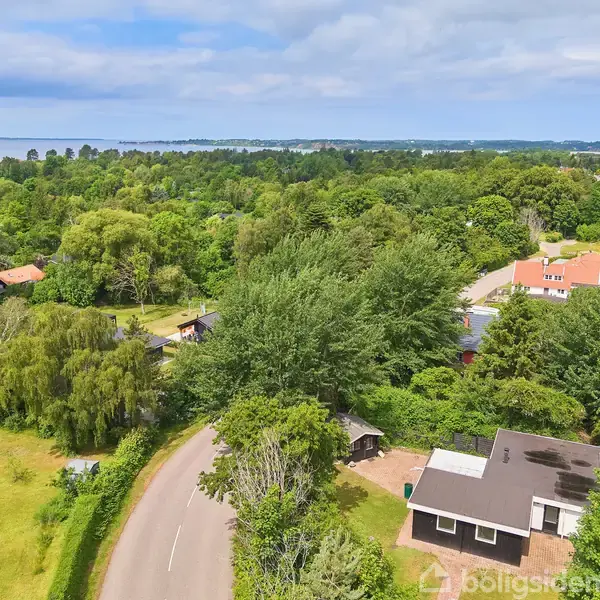 Et hus set fra oven er omgivet af frodige træer og grøn vegetation. En landevej snor sig ved siden af huset i en landlig, skovklædt landskab med spredte bygninger.