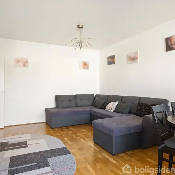 En grå hjørnesofa står i et lyst stuehjørne med hvidmalede vægge dekoreret med små billeder. Et rundt spisebord med stole står ved siden af, og et tæppe dækker parketgulvet.