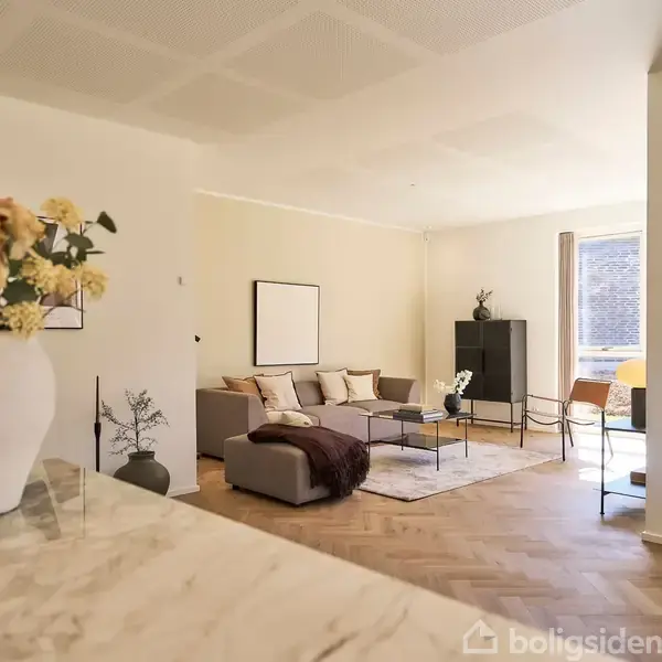 En stue indeholder en sofa med puder og et sofabord foran et stort vindue. En vase med blomster står på et marmorbord. På væggen hænger en plakat med teksten: "SAVE WATER DRINK WINE."