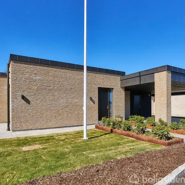 En moderne, kubisk, murstenshus står stille i et beboelsesområde. Huset er omgivet af en velplejet græsplæne og nyplantede blomsterbede under en klar blå himmel.