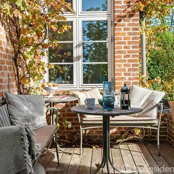 Rundt havebord med kaffepresse og to kopper står på en træterrasse. To stole med puder er placeret ved et teglstenshus med ranker, omgivet af grønne planter.