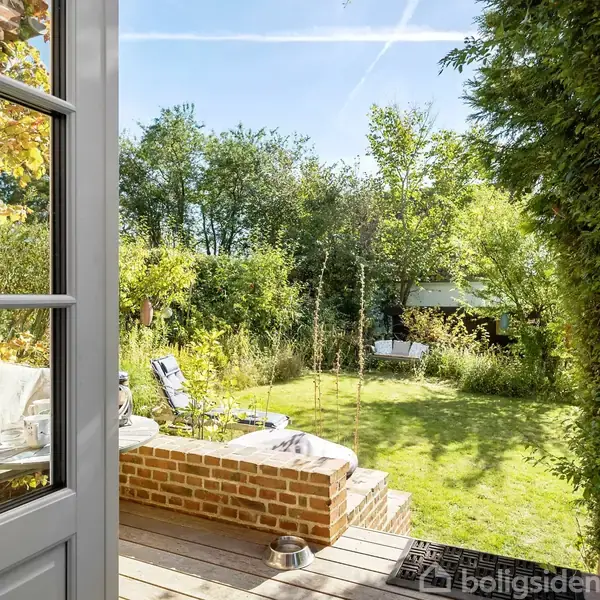 En åben havedør fører ud til en velplejet have med græsplæne, træer og buske. Der er en terrasse med en bænk og puder til afslapning i det solrige vejr.