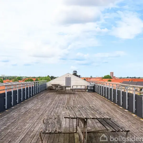 Tagterrasse med træbord i centrum omgivet af rækværk. Skyer strækker sig over lyseblå himmel, mens rødtagede bygninger og grønne træer pryder horisonten i baggrunden.