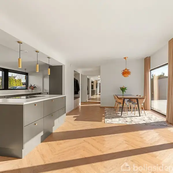 Moderne køkken med grå køkkenø under tre pendellamper, tilstødende spiseområde med spisebord og stole. Store vinduer slipper masser af lys ind, og udsigt til have.