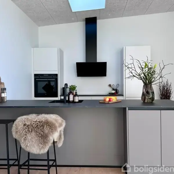 Køkken med sort komfur og hvidt skab, dekoreret med en skindbeklædt stolesæde. En vase med blomster og frugter på bordpladen. Moderne stil, lyst miljø.