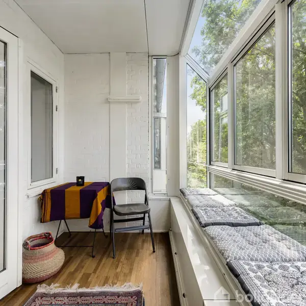 Balkon med en stol ved et bord med farverigt tæppe, omgivet af store vinduer og træer udenfor. Gulvet er af træ, og der er en kurv ved siden af stolen.