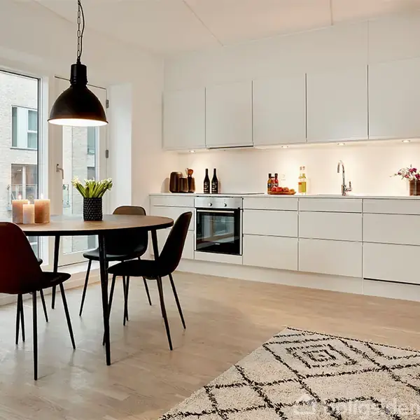 Et moderne køkken med hvide skabe og indbygget ovn. Et rundt spisebord med fire stole står ved store vinduer. Et sort pendellampe hænger over bordet.