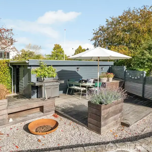 En terrasse med træmøbler og parasol fungerer som en udendørs spiseplads, omgivet af planter i højbede. En grill og bålplads er placeret i en have med grusunderlag.