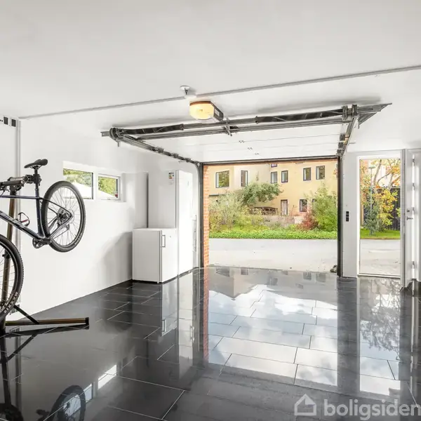 En cykel står på et stativ i en garage med skinnende sorte fliser. Garageporten er åben, og man kan se en bygning udenfor.