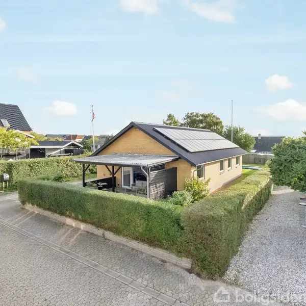 Et gult enfamiliehus med solpaneler på taget står omgivet af en hæk. Huset har en overdækket terrasse mod venstre, og der er grus i indkørslen. Grønt landskab omgiver området.