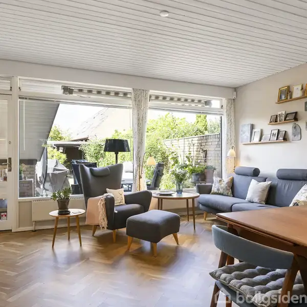 En stue med moderne møbler, herunder en sofa og stol, står foran store vinduer, der åbner ud til en terrasse. Rummet har trægulv og vægdekorationer.