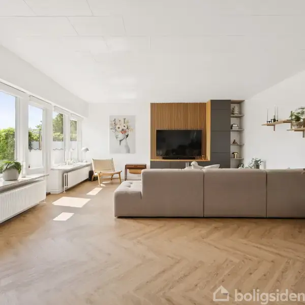 En lys stue har en stor beige sofa foran et TV på et moderne træmøbel. Store vinduer til venstre lukker sollys ind og viser en grøn have udenfor. En potteplante står til højre.