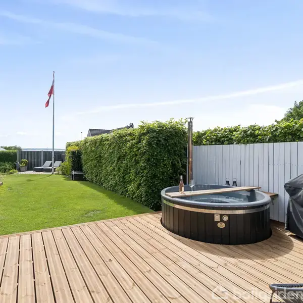 Boblebad står på en træterrasse ved siden af en grill. Det befinder sig i en have med grøn græsplæne og tæt klippet hæk. En flagstang ses i baggrunden.