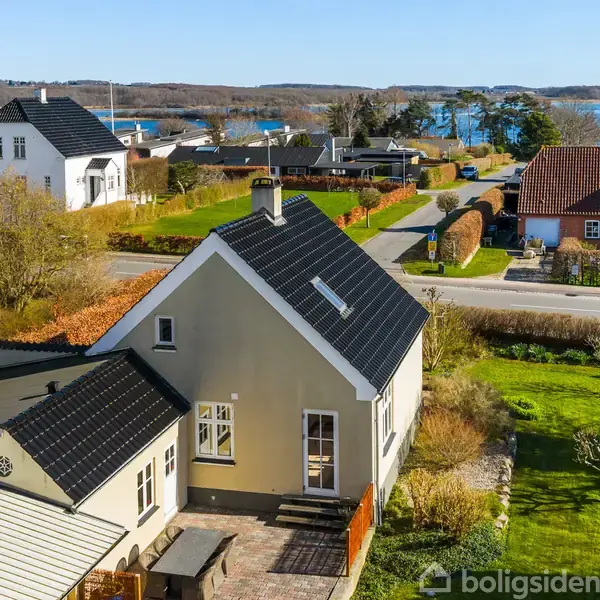 Et hus med sort tag står i en grøn have ved en vej. Omkringliggende huse og en fjord i baggrunden skaber en idyllisk kystnær atmosfære.