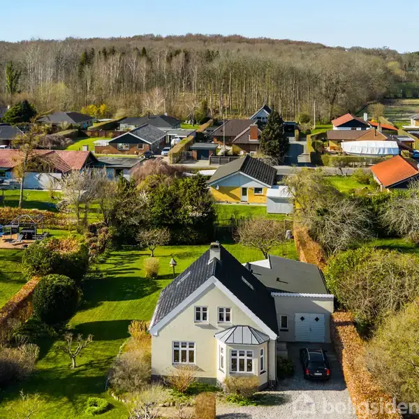 Hus med sort tag og beplantet have står i et villakvarter nær en vej. Kystlinje og blå vand ses i baggrunden.